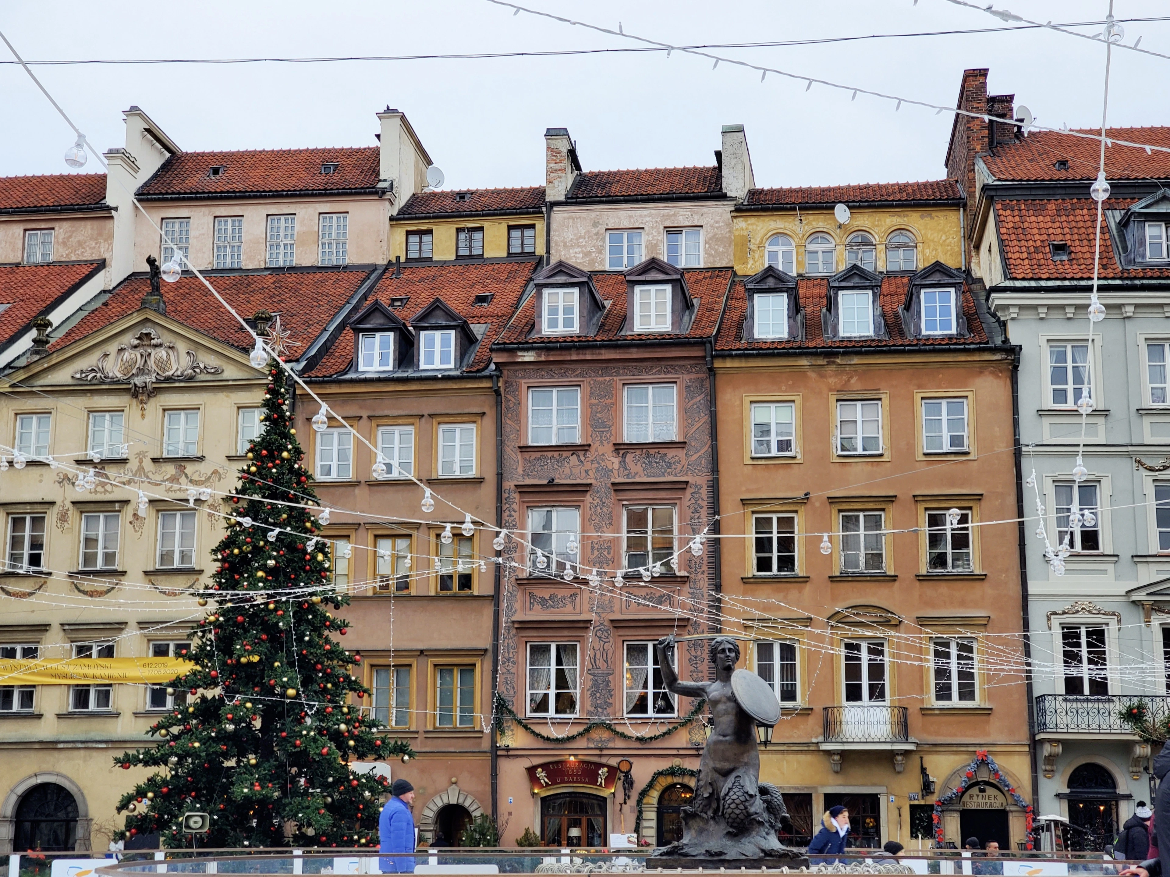 A lit Christmas tree and a fountain statue stand before the colorful townhouses of Warsaw's Old Town Square.