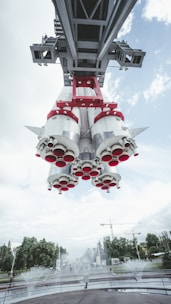 A large rocket suspended above a park-like setting with water fountains below and trees in the background. The view is from underneath the rocket, showcasing its engine nozzles and structural supports. The sky is mostly cloudy with patches of blue visible. Cranes and construction are seen in the distance.