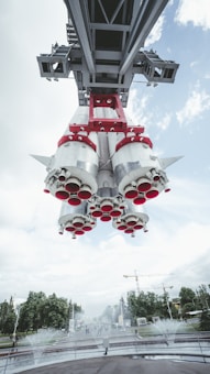 A large rocket suspended above a park-like setting with water fountains below and trees in the background. The view is from underneath the rocket, showcasing its engine nozzles and structural supports. The sky is mostly cloudy with patches of blue visible. Cranes and construction are seen in the distance.