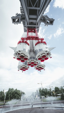 A large rocket suspended above a park-like setting with water fountains below and trees in the background. The view is from underneath the rocket, showcasing its engine nozzles and structural supports. The sky is mostly cloudy with patches of blue visible. Cranes and construction are seen in the distance.