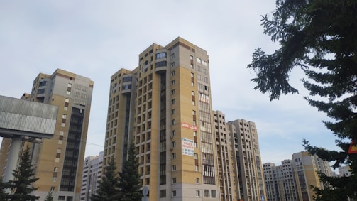 A group of tall residential apartment buildings with modern architecture. The buildings are mostly beige and gray in color and have balconies. There are some pine trees in the foreground, adding greenery to the urban landscape. The sky is clear and blue, suggesting a calm weather day.