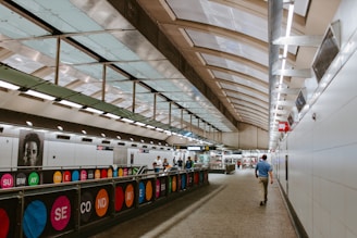 A modern subway station with a sleek, curved ceiling made of glass and metal. The station is well-lit with overhead fluorescent lights and contains a walkway with colorful signage that displays the words 'SUBWAY' and 'SECOND AVENUE'. People are walking through the station, and there is a prominent black-and-white portrait on the wall.