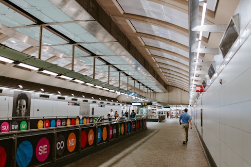 A modern subway station with a sleek, curved ceiling made of glass and metal. The station is well-lit with overhead fluorescent lights and contains a walkway with colorful signage that displays the words 'SUBWAY' and 'SECOND AVENUE'. People are walking through the station, and there is a prominent black-and-white portrait on the wall.