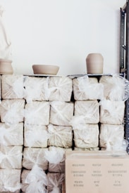 Stacks of moist clay wrapped in plastic bags are arranged on shelves. Two pieces of pottery in the process of being shaped are placed on top. There's a cardboard box in the lower right corner with text in multiple languages.