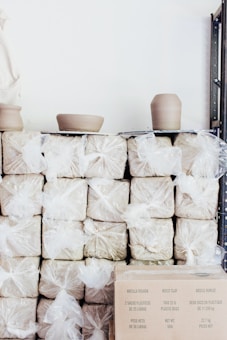 Stacks of moist clay wrapped in plastic bags are arranged on shelves. Two pieces of pottery in the process of being shaped are placed on top. There's a cardboard box in the lower right corner with text in multiple languages.