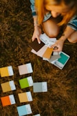 A person holding a selection of Pantone color swatches while kneeling on grassy ground. Several other swatches are spread out on the grass. The person's attire includes a denim jacket, and tattoos are visible on their arms.