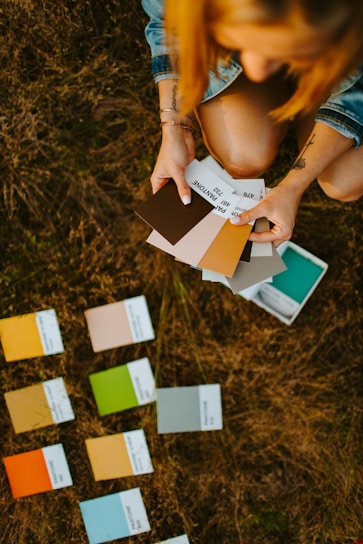 A person holding a selection of Pantone color swatches while kneeling on grassy ground. Several other swatches are spread out on the grass. The person's attire includes a denim jacket, and tattoos are visible on their arms.