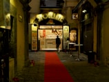 A narrow alleyway leads to a small shop with a prominent sign reading 'Sfogliate'. The shop is lit warmly, displaying pastries inside. A red carpet runs along the cobblestone path towards the shop entrance. Outside, a few small tables are set up, and a person is walking towards the shop.