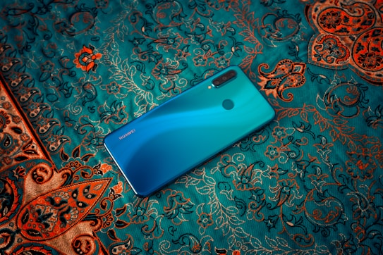 A teal-colored smartphone is placed on an intricately patterned fabric. The fabric features swirling designs in shades of orange and gold against a teal background, creating a luxurious and artistic setting.