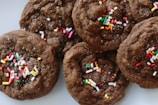 A close-up view of chocolate cookies topped with colorful sprinkles. The cookies appear soft and chewy, with visible granules of sugar and vibrant sprinkles adding a festive touch.