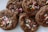 A close-up view of chocolate cookies topped with colorful sprinkles. The cookies appear soft and chewy, with visible granules of sugar and vibrant sprinkles adding a festive touch.