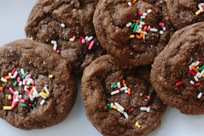 A close-up view of chocolate cookies topped with colorful sprinkles. The cookies appear soft and chewy, with visible granules of sugar and vibrant sprinkles adding a festive touch.