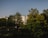 A modern residential building is partially visible behind a group of lush, green trees in a well-maintained park area. The sky is clear and blue, indicating a sunny, pleasant day.