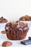 A close-up of a chocolate muffin with a textured, moist surface topped with nuts. The muffin is set against a neutral white background, with chunks of chocolate and pecans nearby. Two additional muffins are visible in the blurred background.