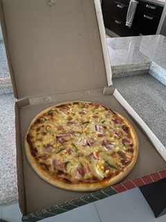A freshly baked pizza inside an open cardboard box resting on a marble countertop. The pizza is topped with ingredients like ham, green peppers, corn, and cheese, creating a colorful appearance.