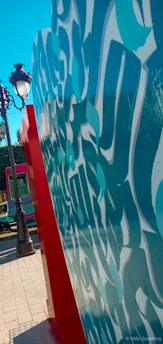 A large piece of modern urban art features abstract turquoise and white patterns on a vertical surface, set in an outdoor urban environment. The artwork is bordered by a bright red edge. In the background, a classic streetlamp and a tram are visible, along with some trees and paving stones.