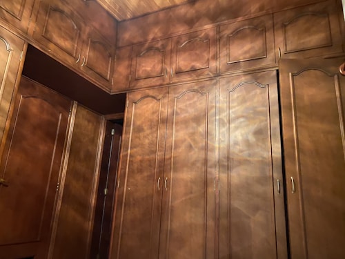 A view of tall, wooden cabinets filling the walls of a room. The cabinets have ornate borders on the doors and brass handles. The wood has a warm, rich color with light reflections creating a shimmering effect. Part of the doorway is also visible.