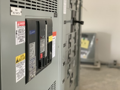 A close-up view of an industrial electrical panel with various switches and warning labels. The panel features caution and danger signs in multiple languages, and its metallic surface is complemented by the soft lighting of the room.