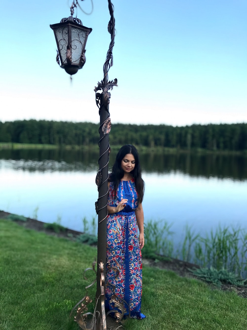 Mujer en vestido floral azul y rosa de pie en el campo de hierba verde cerca del lago durante el día