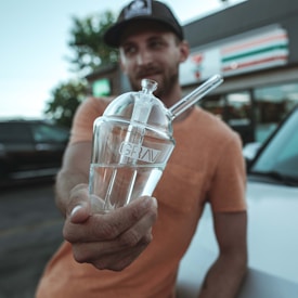 A person in a casual orange t-shirt and a dark cap is holding a clear glass smoking device marked 'GRAV' outside a convenience store. The store and a parked vehicle are blurred in the background.