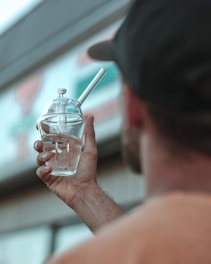 A person holds up a glass object with water inside, featuring a logo that reads 'GRAV'. The background is blurred, showing part of a sign or building.