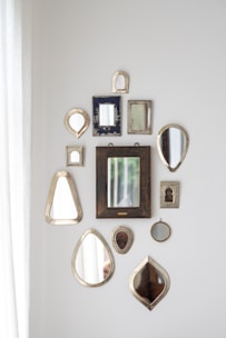 A collection of various decorative mirrors arranged on a white wall in different shapes and sizes. The mirrors have ornate or simple frames with a mix of metallic, wooden, and textured finishes. Natural light enters from the left, adding a soft illumination to the arrangement.