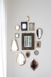 A collection of various decorative mirrors arranged on a white wall in different shapes and sizes. The mirrors have ornate or simple frames with a mix of metallic, wooden, and textured finishes. Natural light enters from the left, adding a soft illumination to the arrangement.