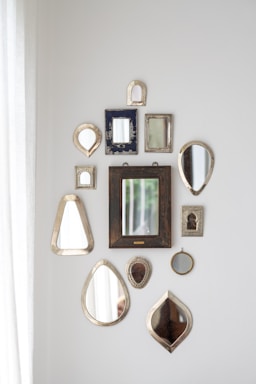 A collection of various decorative mirrors arranged on a white wall in different shapes and sizes. The mirrors have ornate or simple frames with a mix of metallic, wooden, and textured finishes. Natural light enters from the left, adding a soft illumination to the arrangement.