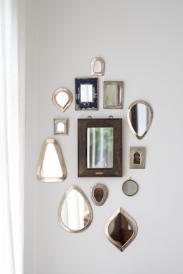 A collection of various decorative mirrors arranged on a white wall in different shapes and sizes. The mirrors have ornate or simple frames with a mix of metallic, wooden, and textured finishes. Natural light enters from the left, adding a soft illumination to the arrangement.