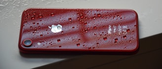 A red smartphone with water droplets on its surface rests on a light-colored surface, emphasizing its waterproof feature and sleek design.