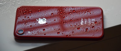 A red smartphone with water droplets on its surface rests on a light-colored surface, emphasizing its waterproof feature and sleek design.