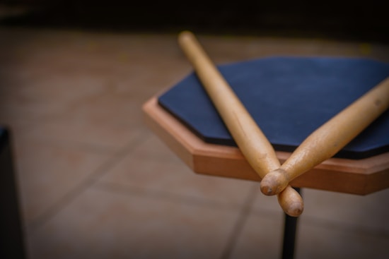 A pair of wooden drumsticks rests on a practice pad with a textured surface. The pad has a black hexagonal striking area and a wooden border, set on a stand against a blurred background.