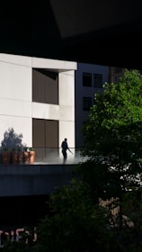 A person is standing on an outdoor balcony or terrace of a modern building, surrounded by a glass railing. The building has large windows and a sleek design. There are potted plants along the edge of the balcony and green foliage in the foreground, casting shadows on the wall.
