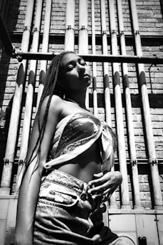 A person stands confidently against an industrial backdrop with cylindrical pipes and a textured brick wall. The person is wearing jeans and a patterned top, and the lighting creates a bold contrast.