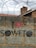 A brick wall topped with barbed wire features graffiti. The text 'I ♥ SOWETO' is painted on the wall. In the background, there are tiled roofs and an antenna.