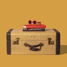 A vintage suitcase is placed against a solid mustard yellow background. On top of the suitcase, there are two books stacked, the top one being red. A pair of red, round sunglasses rest on the books.