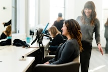A woman sits in a salon chair with a smile on her face as a stylist, also smiling, stands beside her. Various salon tools including a hair dryer and brush are visible on the counter. The room is well-lit with large windows in the background, creating a bright and cheerful atmosphere.