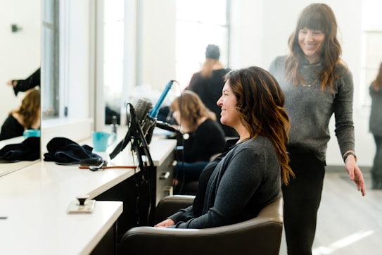 A woman sits in a salon chair with a smile on her face as a stylist, also smiling, stands beside her. Various salon tools including a hair dryer and brush are visible on the counter. The room is well-lit with large windows in the background, creating a bright and cheerful atmosphere.