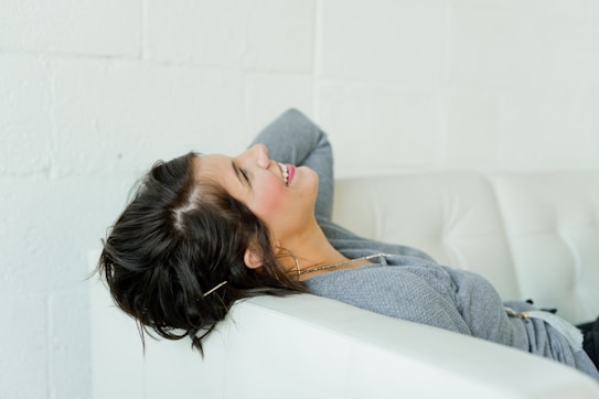 A woman is leaning back comfortably on a white couch, with her head tilted back and a relaxed smile on her face. She is wearing a gray sweater and appears to be enjoying a moment of relaxation.