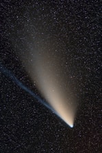 A bright comet with a distinctive glowing tail streaks across a star-filled night sky. The sky is densely populated with small, twinkling stars, creating a contrasting backdrop for the comet's luminous presence.