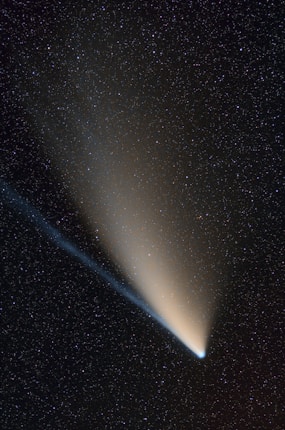 A bright comet with a distinctive glowing tail streaks across a star-filled night sky. The sky is densely populated with small, twinkling stars, creating a contrasting backdrop for the comet's luminous presence.