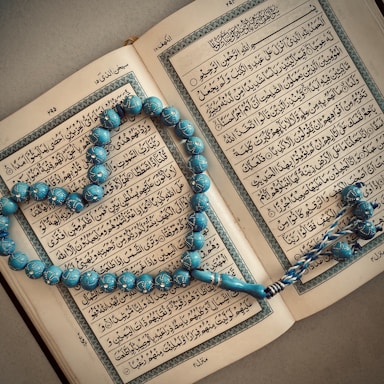 An open book with Arabic script is displayed, with a string of blue prayer beads arranged in a heart shape on top. The pages are richly decorated with borders, and the beads have intricate patterns.