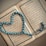 An open book with Arabic script is displayed, with a string of blue prayer beads arranged in a heart shape on top. The pages are richly decorated with borders, and the beads have intricate patterns.
