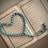 An open book with Arabic script is displayed, with a string of blue prayer beads arranged in a heart shape on top. The pages are richly decorated with borders, and the beads have intricate patterns.