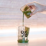A hand wearing a transparent glove is pouring a green liquid from a small glass container into a clear plastic cup filled with ice cubes. The cup has a percentage symbol printed on it. The background is softly lit with a warm tone.
