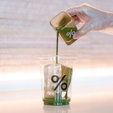 A hand wearing a transparent glove is pouring a green liquid from a small glass container into a clear plastic cup filled with ice cubes. The cup has a percentage symbol printed on it. The background is softly lit with a warm tone.