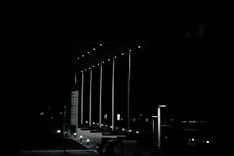 A nighttime scene with minimal lighting, featuring tall, slender poles with small lights near the top. These poles are part of a modern architectural structure with a flat roof. Dimly lit bollards line a pathway leading into the darkness.