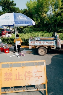 A street vendor sits under a blue and white China Taiping umbrella, wearing a mask and a yellow shirt. Beside them is a tricycle with Chinese characters written on it. Nearby, there are red buckets filled with various items. The scene is set in a leafy area with a no-parking sign in the foreground.