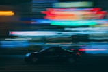 A dynamic scene with a car speeding through an urban setting. Brightly colored neon lights create a blur of motion in the background, emphasizing movement and city nightlife.