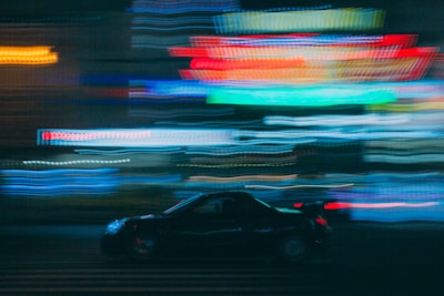 A dynamic scene with a car speeding through an urban setting. Brightly colored neon lights create a blur of motion in the background, emphasizing movement and city nightlife.